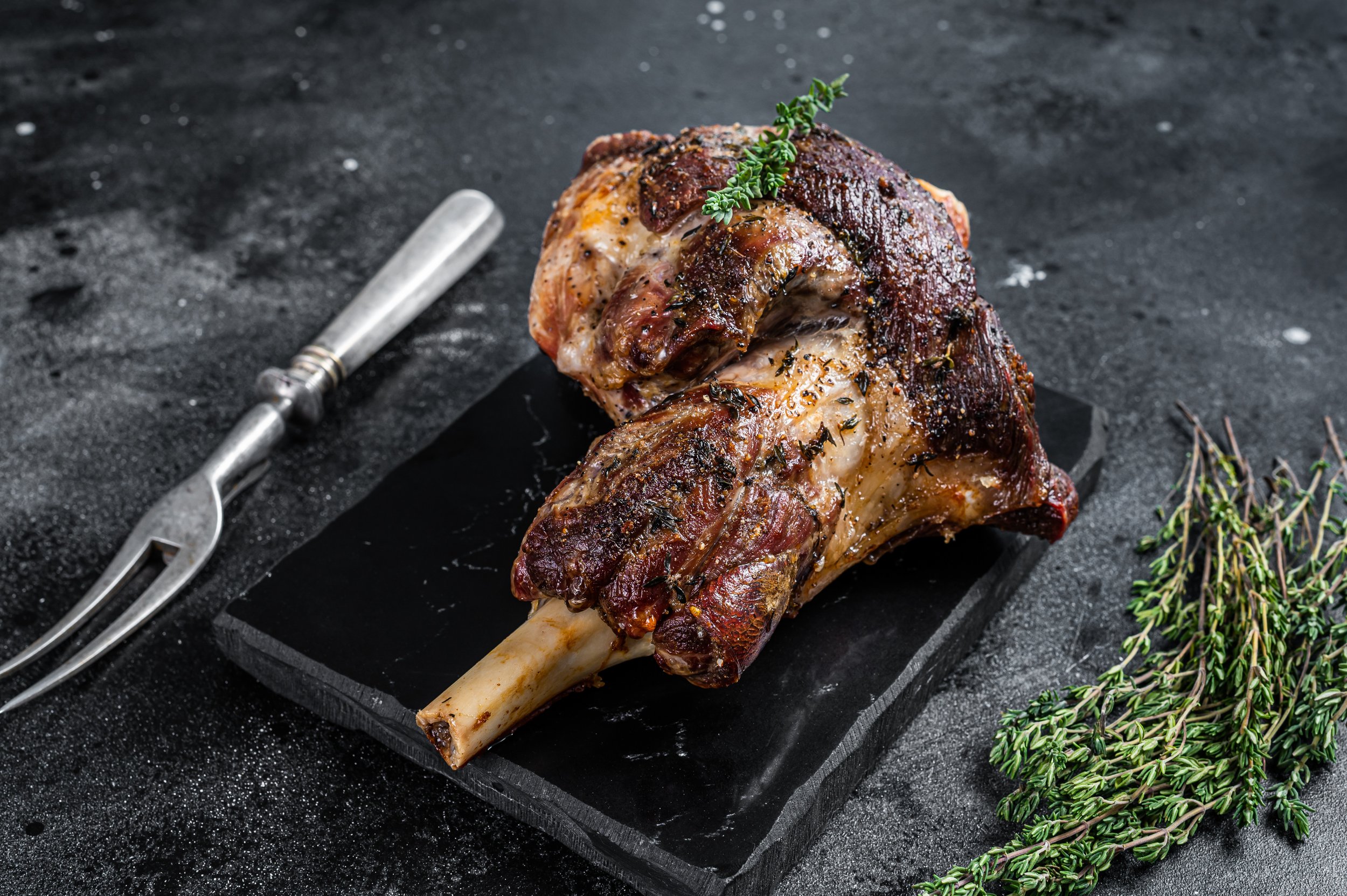 Roasted lamb shank garnished with fresh thyme on a black slate serving board, accompanied by a metal carving fork and sprigs of thyme on a dark textured background.