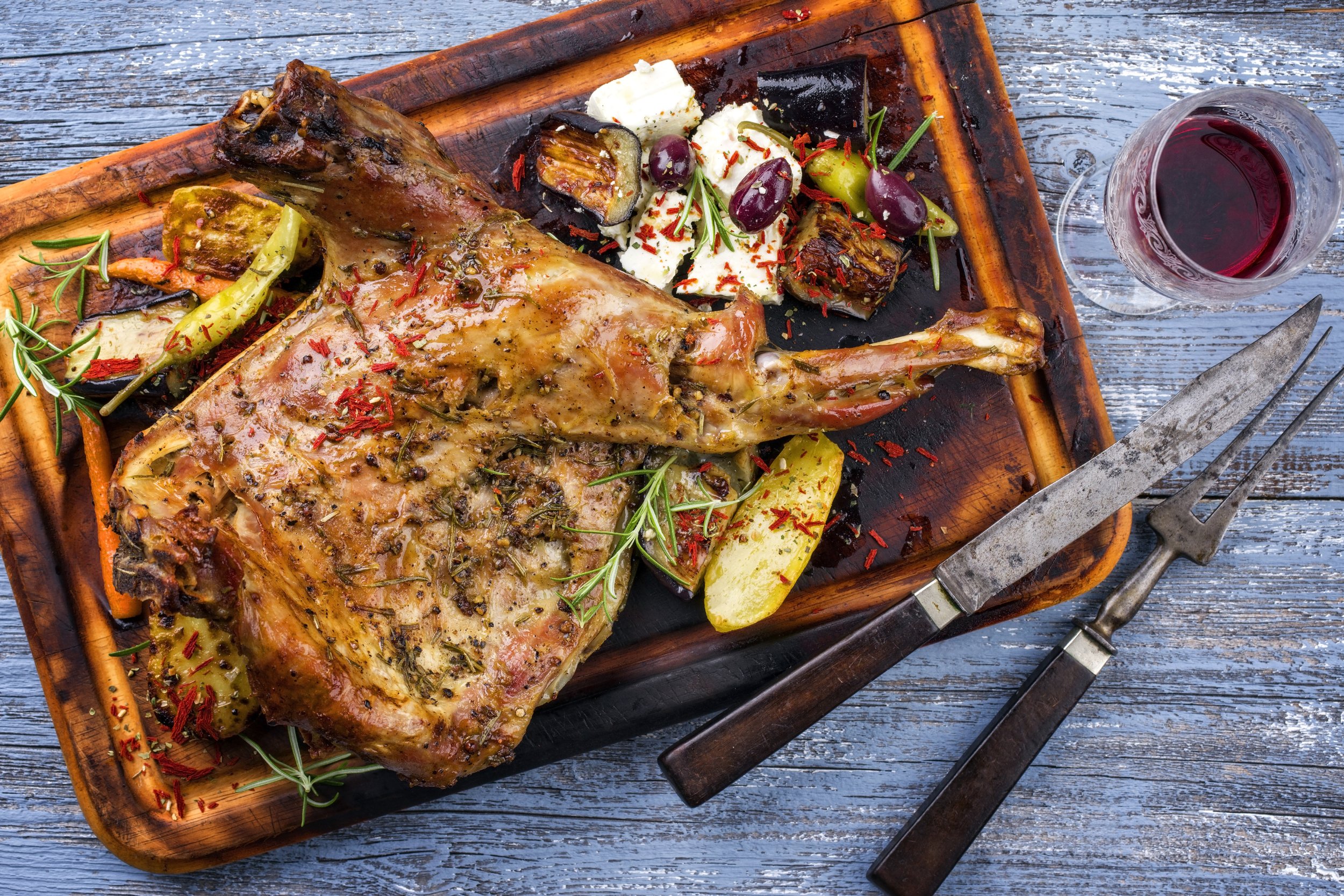 Roasted leg of lamb garnished with herbs, surrounded by grilled vegetables and olives, served on a wooden platter with a glass of red wine and silver cutlery.
