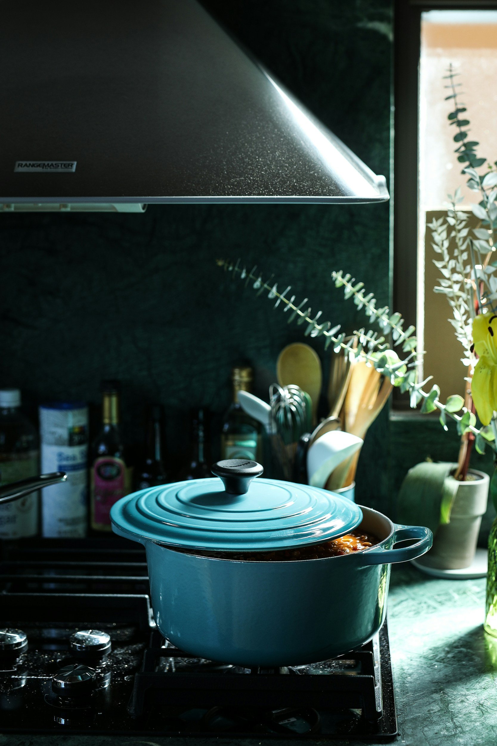 A turquoise cooking pot on a gas stove in a modern kitchen, surrounded by cooking utensils and bottles. The scene features a dark green wall and natural light illuminating the space, creating a warm and inviting atmosphere.